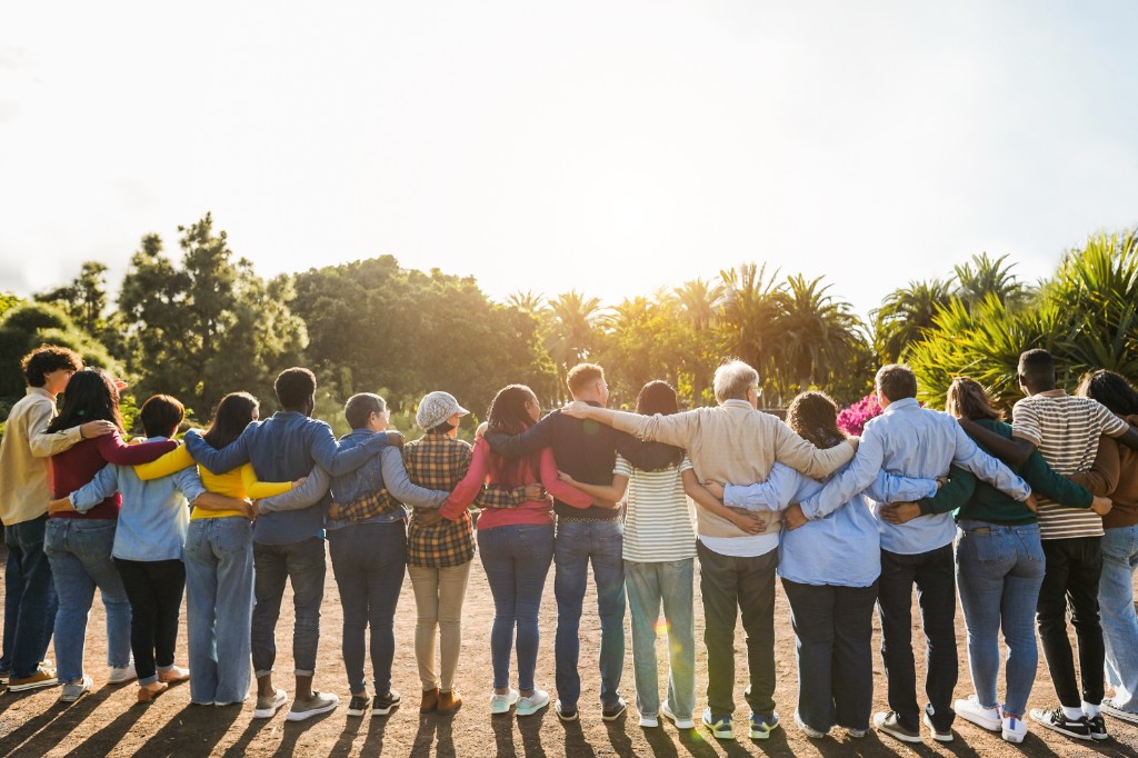 A diverse group of people standing together outdoors in warm evening light, seen from behind with their arms around one another.